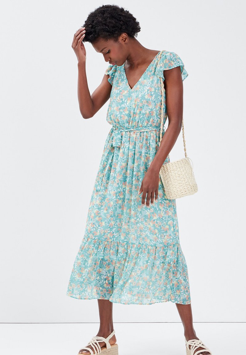 Robe midi florale en turquoise clair avec un décolleté en V, des manches courtes à volants et une taille cintrée. Présente un ourlet à volants et un sac en osier.