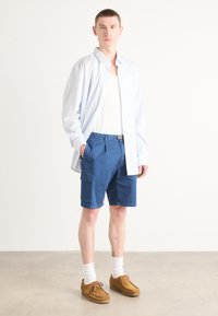 Chemise boutonnée rayée bleu clair, débardeur blanc, shorts cargo bleus, chaussures en cuir marron à lacets, chaussettes blanches, debout sur un fond neutre.