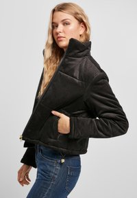 Zwarte corduroy jas met een hoge kraag, gewatteerd ontwerp en ritsluiting. Heeft een geribbelde textuur en zijzakken. Gepaard met blauwe jeans.