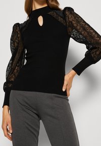 Haut noir côtelé avec des manches bouffantes en dentelle, col haut et détail en trou de serrure. Silhouette ajustée avec des accents texturés et une finition lisse.