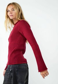 Rotes ribbeltes Langarmoberteil mit Rundhalsausschnitt und ausgestellten Ärmelabschlüssen, kombiniert mit dunklen Jeans. Die Textur ist ausgeprägt und die Silhouette betont die Figur.