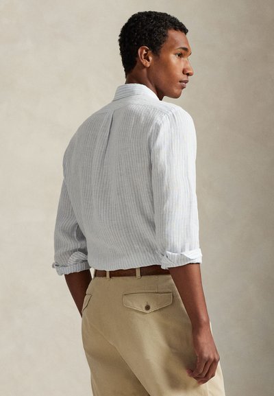 Ανοιχτό μπλε πουκάμισο με ρίγες και μακριά μανίκια, τυλιγμένα προς τα πάνω, συνδυασμένο με χακί παντελόνι που έχει πίσω τσέπες και καφέ ζώνη.