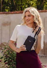 Black leather crossbody bag with a zippered pocket, featuring a logo on the front. Model is wearing a white top and burgundy skirt.