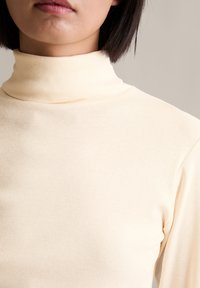 Cremfarbener Rollkragenpullover mit geripptem Textur, enganliegendem Design und hohem Kragen. Der Stoff wirkt weich und elastisch mit nahtlosen Schultern.