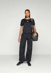 Zwart sportoutfit bestaande uit een shirt met korte mouwen en losse broek, beide met het Adidas-logo. Een kleine zwarte tas hangt over de schouder.