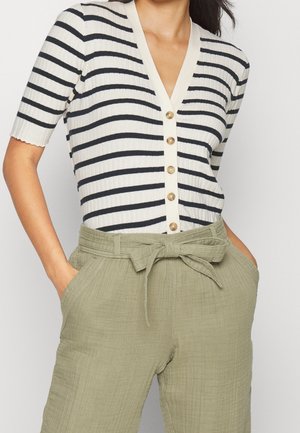 Femme portant une chemise à manches courtes rayée noir et blanc, boutonnée, rentrée dans un pantalon taille haute couleur olive clair avec ceinture nouée et mains dans les poches.