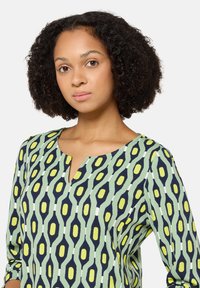 Grüne Bluse mit marineblauem und gelbem abstraktem Muster, mit V-Ausschnitt und dreiviertellangen Ärmeln, aus leichtem Stoff gefertigt.