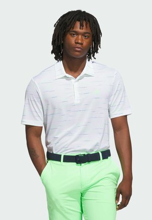 Junger Mann mit Dreadlocks, der ein weißes kurzärmeliges Polohemd und leuchtend grüne Hosen mit einem schwarzen gewebten Gürtel trägt, steht mit den Händen in den Taschen.
