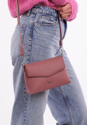 Žena v modrých džínech a růžovém svetru drží malou crossbody tašku ve dusty rose barvě se stříbrným klopovým logo. Na prstu je viditelný prsten.