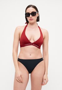 Rød haltert bikini topp med dyp V-hals og logo-bånd, paret med svarte bikini-bunner med sidesnitt og glatt tekstur.