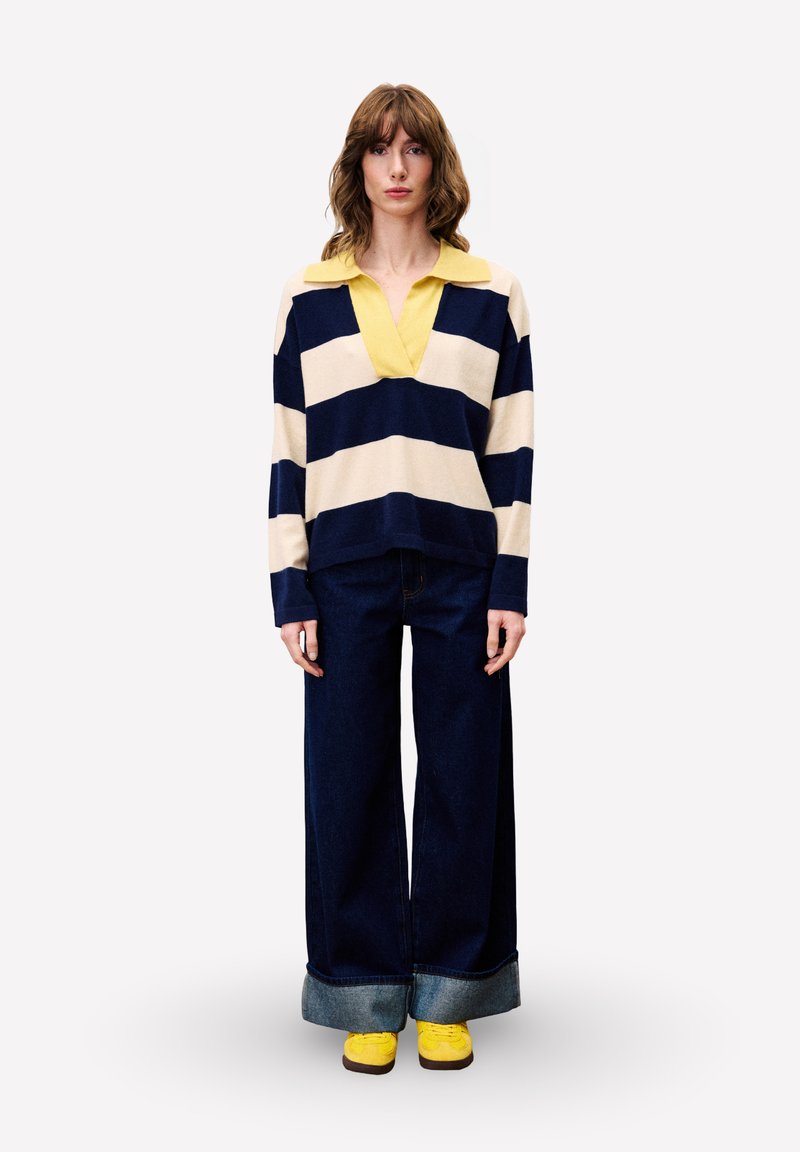 Vrouw staat op en draagt een gestreepte lange mouwen shirt in marineblauw en roomkleur met een gele kraag, wijde donkere jeans opgerold aan de onderkant, en gele schoenen.