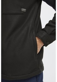 Czarny polarowy sweter z przednią kieszenią, detalami przy mankietach na guziki, gładką teksturą i metką z logo na kieszeni. Prosty, funkcjonalny design.