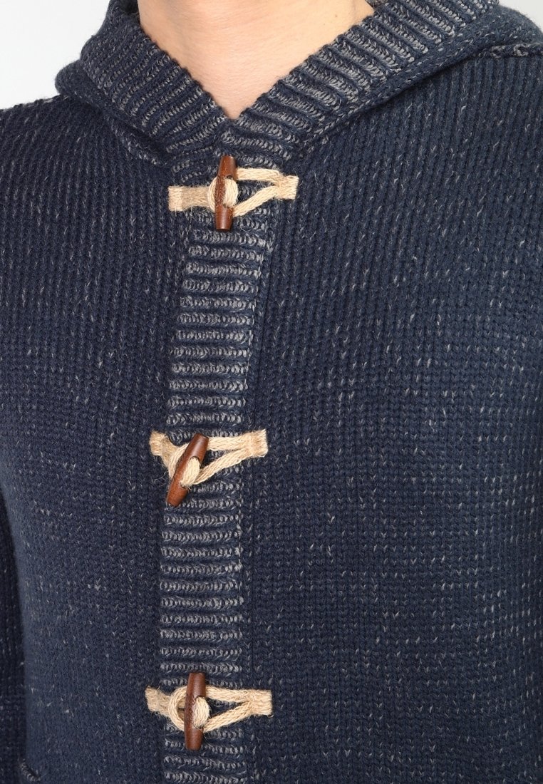Mörkblå stickad hoodie med texturerad tyg, inbyggda träspännen, beige detaljer och ett ribbat mönster längs öppningen.