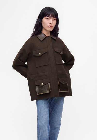 Jeune femme aux longs cheveux noirs porte une veste marron foncé avec de grandes poches et un col en cuir, associée à un jean bleu, debout sur un fond blanc.