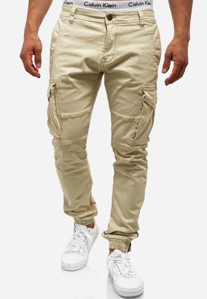 Pantalon cargo beige avec plusieurs poches et poignets élastiques, porté avec des baskets blanches et une ceinture Calvin Klein visible.