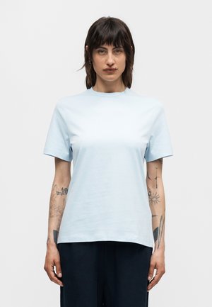 Γυναίκα με σκούρα μαλλιά μέχρι τους ώμους, που φοράει ένα ανοιχτό μπλε κοντομάνικο μπλουζάκι και μαύρο παντελόνι, στέκεται μπροστά σε ένα απλό λευκό φόντο.