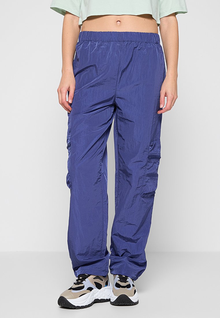 Blaue Cargo-Hosen mit einer leichten, glänzenden Textur, ausgestattet mit einem elastischen Taillenbund und seitlichen Taschen, kombiniert mit gemusterten Turnschuhen.