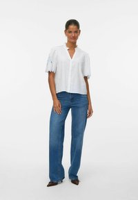 Blusa bianca a maniche corte con bordi ondulati e ricamo azzurro chiaro, abbinata a jeans in denim blu a vita alta e scarpe con tacco marrone scuro.
