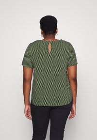 Kvinna med kort lockigt hår iförd en grön och svart mönstrad kortärmad topp och svarta byxor, vänd bort mot en enfärgad bakgrund.