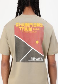 Bežinis marškinėliai su grafiniu dizainu ant nugaros, kuriame raudonos, juodos ir geltonos spalvos. Tekstas sako "CHAMPIONS TRAIN" ir "REPLAY."