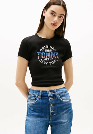 Top corto a maniche corte nero con la scritta "TOMMY" in blu e rosso con stelle, abbinato a jeans strappati a vita alta di un blu chiaro.