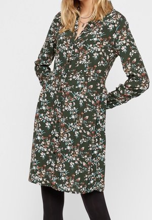 Kobieta ubrana w ciemnozieloną, długą sukienkę z długim rękawem do kolan z wielokolorowym kwiatowym wzorem oraz czarne rajstopy.