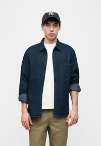 Νεαρός άνδρας που φοράει μπλε πουκάμισο με κουμπιά πάνω από λευκό μπλουζάκι, χακί παντελόνι και μαύρο-άσπρο καπέλο μπέιζμπολ με ρίγες, στέκεται μπροστά σε λευκό φόντο.