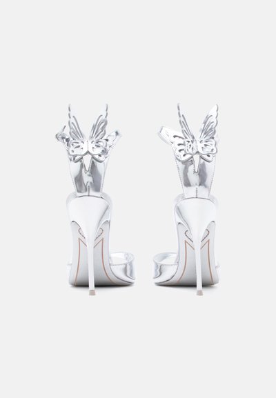 Ασημένια σανδάλια με λεπτά τακούνια στιλέτο και διακοσμήσεις σε σχήμα πεταλούδας στα λουριά του αστραγάλου, εμφανιζόμενα από πίσω.