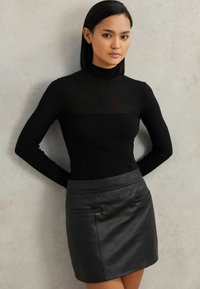 Eine Frau steht gegen eine graue Wand und trägt einen schwarzen Rollkragenpullover sowie einen schwarzen Leder-Minirock mit Taschendetails.