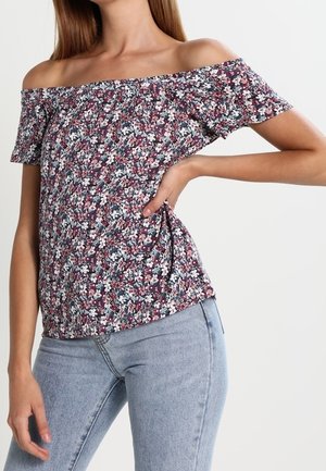 Kvinde iført off-shoulder blomstret top med lilla, hvide og pink blomster, kombineret med lyseblå jeans, hånd hvilende på hoften.