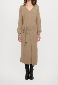 Robe en maille couleur camel avec un col en V, des manches longues et une ceinture à la taille. Présente un design cache-cœur et des accents côtelés aux poignets et à l'ourlet. Bottes noires montantes jusqu'au genou.