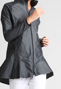 Chaqueta negra, ligera, con cremallera y un alto cuello, textura perforada, dobladillo acampanado y mangas alargadas, diseñada para la comodidad y el movimiento.