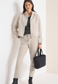 Veste beige avec capuche, taille élastique, associée à un pantalon beige assorti avec poches. Sac noir avec poignées tenu dans la main droite.