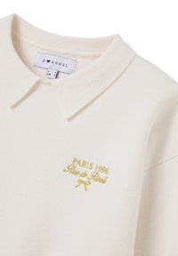 Weißer Polo-Pullover mit strukturiertem Stoff, klassischem Kragen und gesticktem goldenen Schriftzug "PARIS 1996 Rue de Rivoli" sowie einer Schleifendetail.