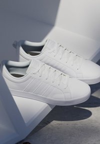 Par de zapatillas bajas blancas limpias con cordones descansando sobre una superficie geométrica blanca que proyecta sombras sobre un suelo gris.