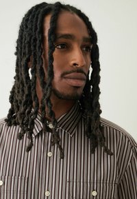 Męska brązowo-kremowa koszula w paski z kołnierzykiem zapinanym na guziki, dwoma kieszeniami na piersi i teksturowanym materiałem. Posiada luźny krój i widoczne guziki.