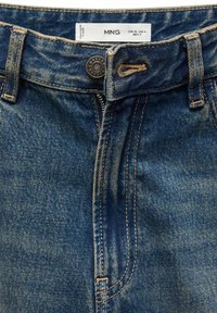 Gros plan sur un jean en denim bleu avec fermeture par bouton, braguette zippée, passants de ceinture et étiquette de marque visible à l’intérieur de la ceinture.