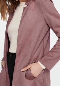 Hellrosa jacke aus wildlederähnlichem Material mit Reverskragen und Seitentaschen, die eine glatte Textur und eine taillierte Passform aufweist, getragen über einem cremefarbenen Oberteil.