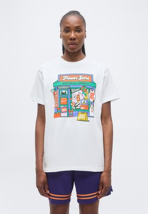 Kvinde iført en hvid T-shirt med farverig blomsterbutiksgrafik og lilla shorts med orange striber på en ensfarvet hvid baggrund.