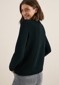 Donkergroene longsleeve met een ronde hals, gladde textuur en een ontspannen pasvorm. Gekleed met lichtgrijze jeans, wat een casual stijl laat zien.