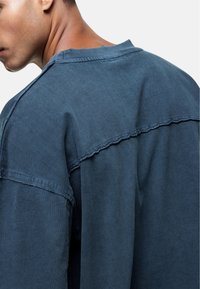 Chemise en coton bleue oversize avec un dos structuré, des coutures en évidence et un col brut. Texture lisse avec une coupe décontractée.