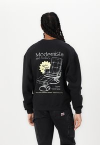 Fekete pulóver, amelyen egy középkori lounger szék grafikája látható, a hátsó részen pedig "Modernista" és "Relaxálj stílusosan" feliratokkal.