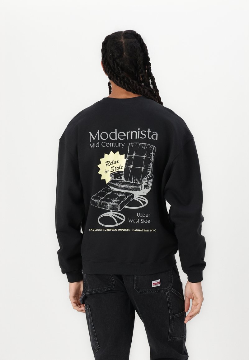 Fekete pulóver, amelyen egy középkori lounger szék grafikája látható, a hátsó részen pedig "Modernista" és "Relaxálj stílusosan" feliratokkal.