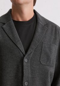 Grijze, gestructureerde wollen blazer met een ingesneden kraag, drie knopen en een enkele borstzak. Draagt een zwart overhemd eronder.