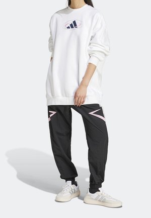 Vit oversizad sweatshirt med grafisk logotyp, kombinerad med svarta joggerbyxor med rosa detaljer och elastiska muddar, kompletterad med vita sneakers.