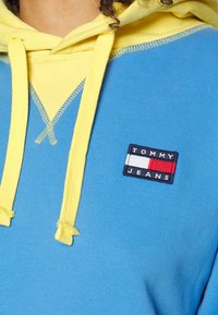 Sudadera con capucha que presenta una tela de contraste azul y amarilla, textura acanalada y un parche con el logo de Tommy Jeans en el pecho.