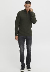 Olivgrüner Pullover mit hohem Kragen und strukturiertem Strick, kombiniert mit dunkelgrauen, enganliegenden Jeans und schwarzen Sneakers. Figurbetonte Silhouette, minimales Design.