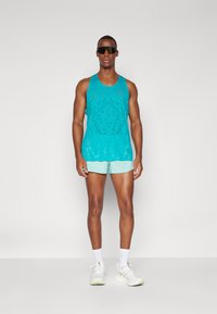 Un débardeur sans manches turquoise avec un motif texturé, associé à un short vert menthe clair. Le mannequin porte des lunettes de soleil noires et des chaussures de sport blanches.
