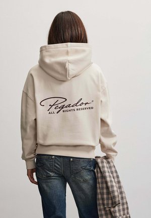 Person med langt brunt hår iført beige hættetrøje med teksten "Pegador All Rights Reserved" på ryggen, holder en foldet ternet jakke i hånden.