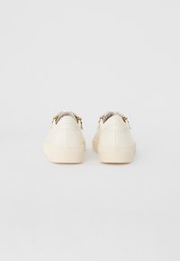 Zapatillas de cuero color crema con un acabado suave, que presentan una suela gruesa y texturizada, así como un detalle decorativo de hardware dorado en la parte trasera.